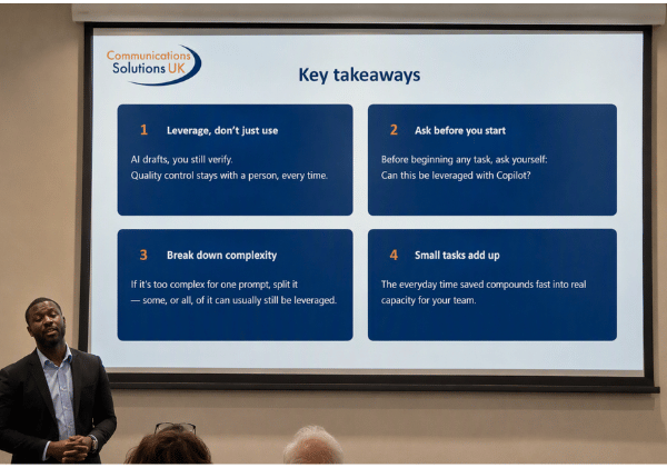 Presentation slide titled “Key takeaways” displayed on a projection screen, featuring the Communications Solutions UK logo and four key points about using AI and Copilot effectively. The points are: (1) “Leverage, don’t just use” with the message that AI drafts but people must verify; (2) “Ask before you start” encouraging users to consider whether a task can be leveraged with Copilot; (3) “Break down complexity” by splitting complex tasks into smaller prompts; and (4) “Small tasks add up” highlighting that everyday time savings create meaningful capacity gains. A presenter stands at the front left of the room, with audience members seated in the foreground.